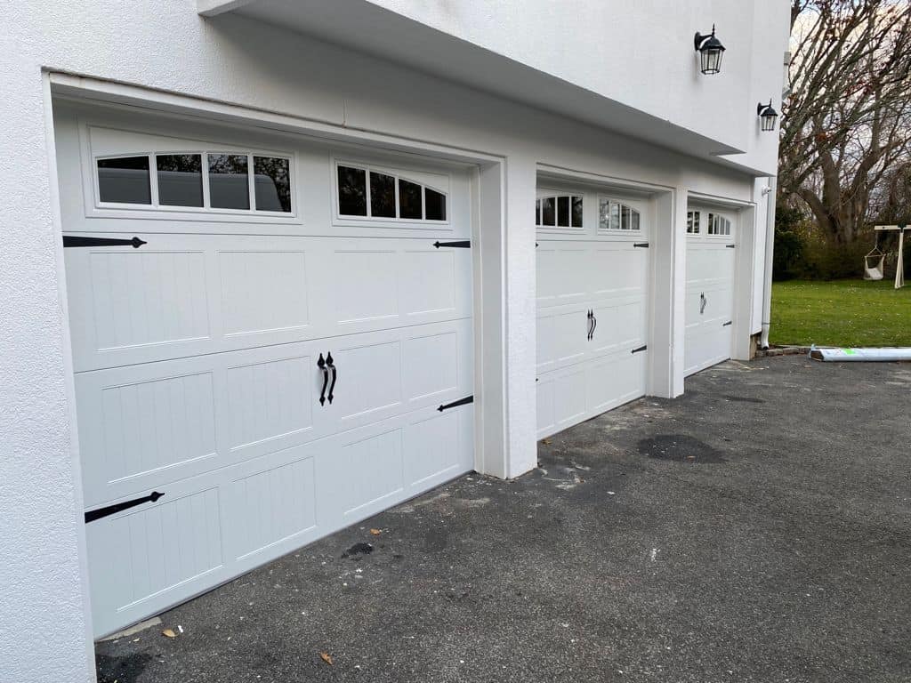 Three white garage doors with black handles and decorative hinges on a white building, next to a paved driveway. Grass and trees are visible in the background—highlighting affordable garage door solutions for any home.