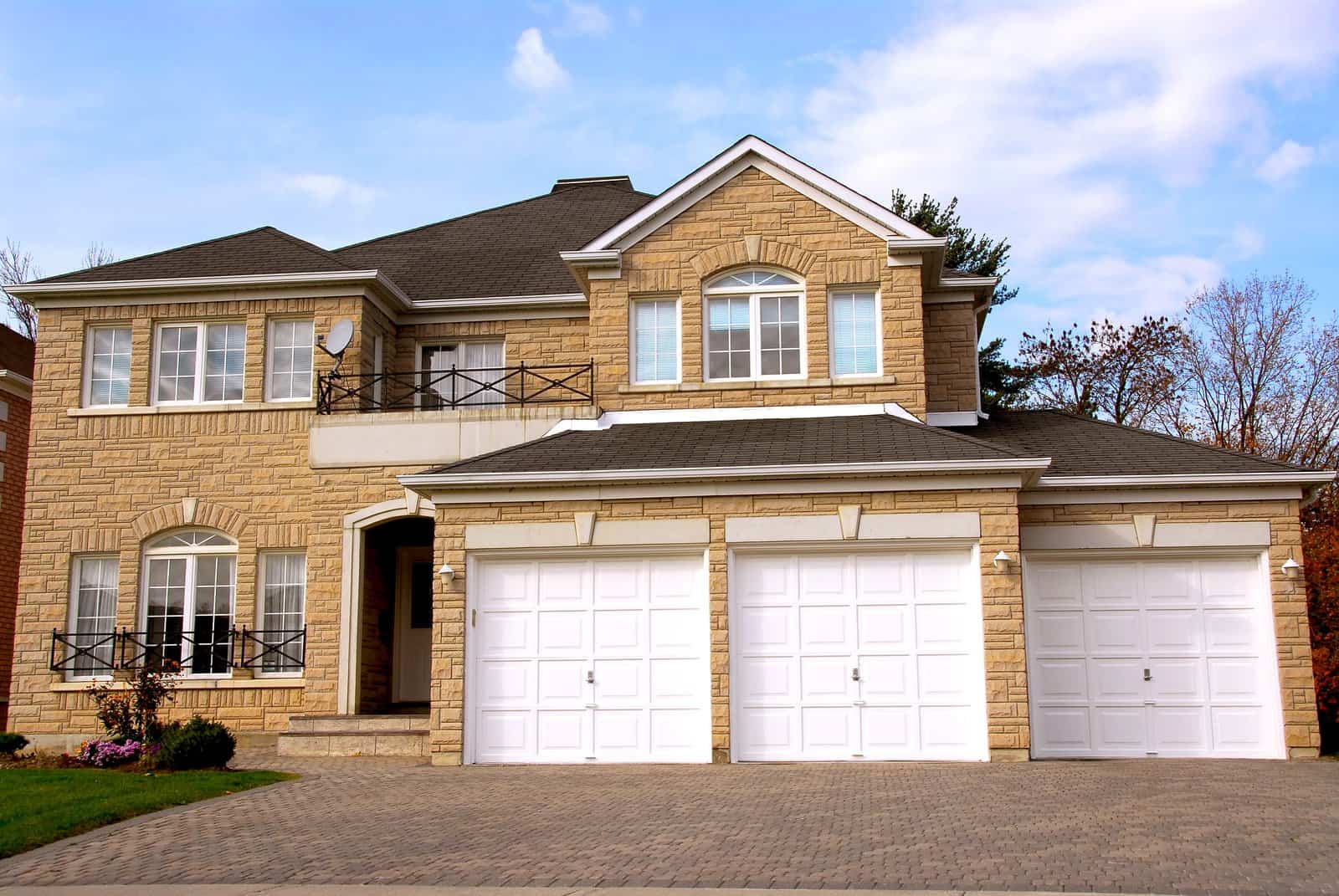 Two-story house with beige brick exterior, black railings, white trim, and three white garage doors—showcasing affordable garage door solutions. The brick-paved driveway is bordered by trees and shrubs in the background.