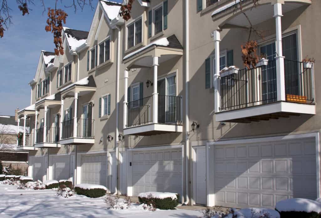 Row of modern townhouse buildings with balconies and white garage doors featuring durable garage openers, set on a snowy street with patches of snow on the ground and some bare trees visible.