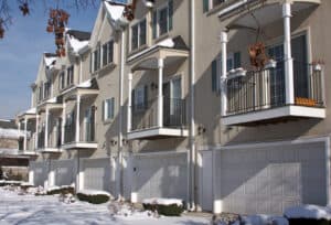 Row of modern townhouse buildings with balconies and white garage doors featuring durable garage openers, set on a snowy street with patches of snow on the ground and some bare trees visible.