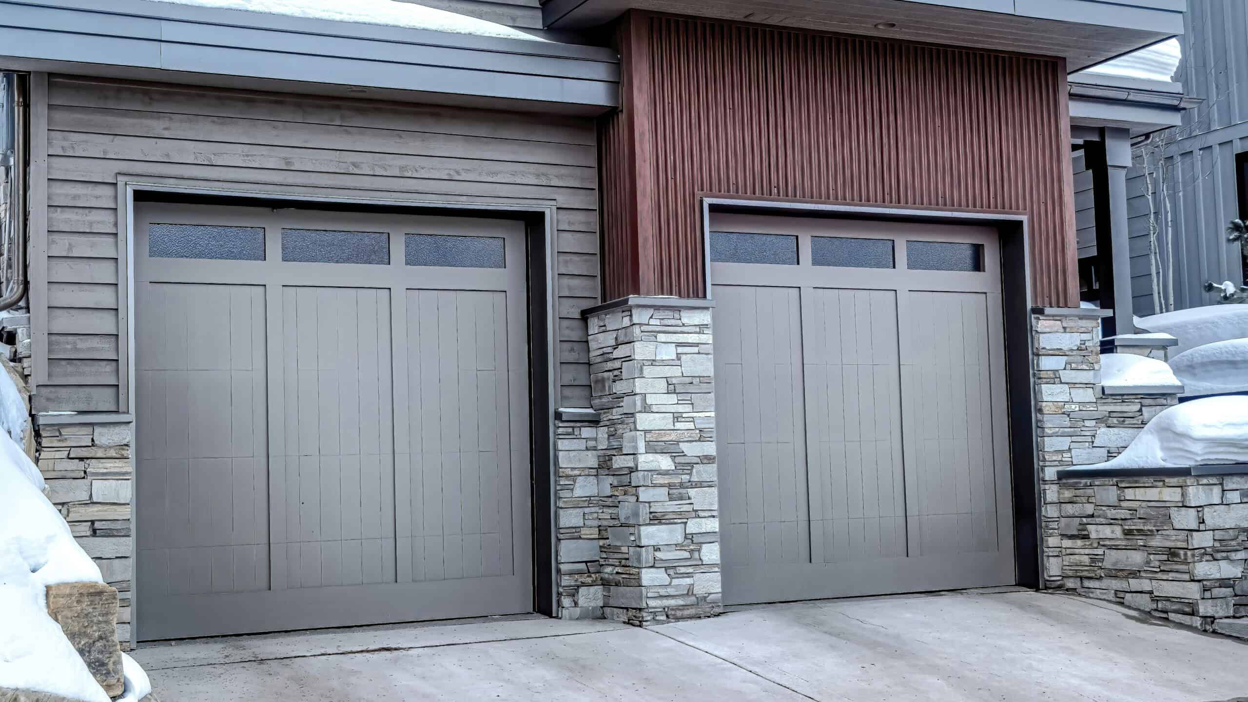 Two closed garage doors with rectangular windows on a modern house exterior featuring wood and stone finishes, surrounded by snow—showcasing affordable garage door solutions built for both style and durability.