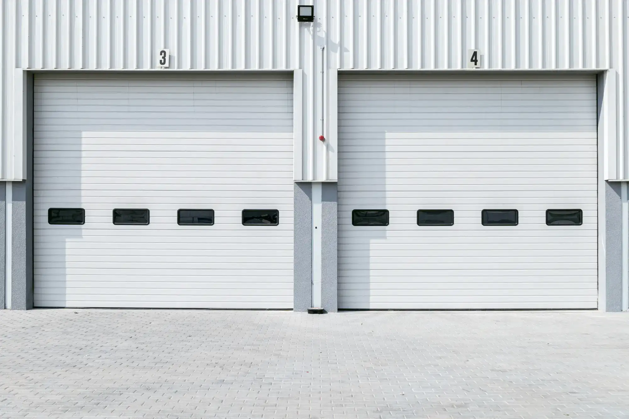 Two closed white industrial garage doors numbered 3 and 4, set in a metal warehouse facade with small rectangular windows on each door.
