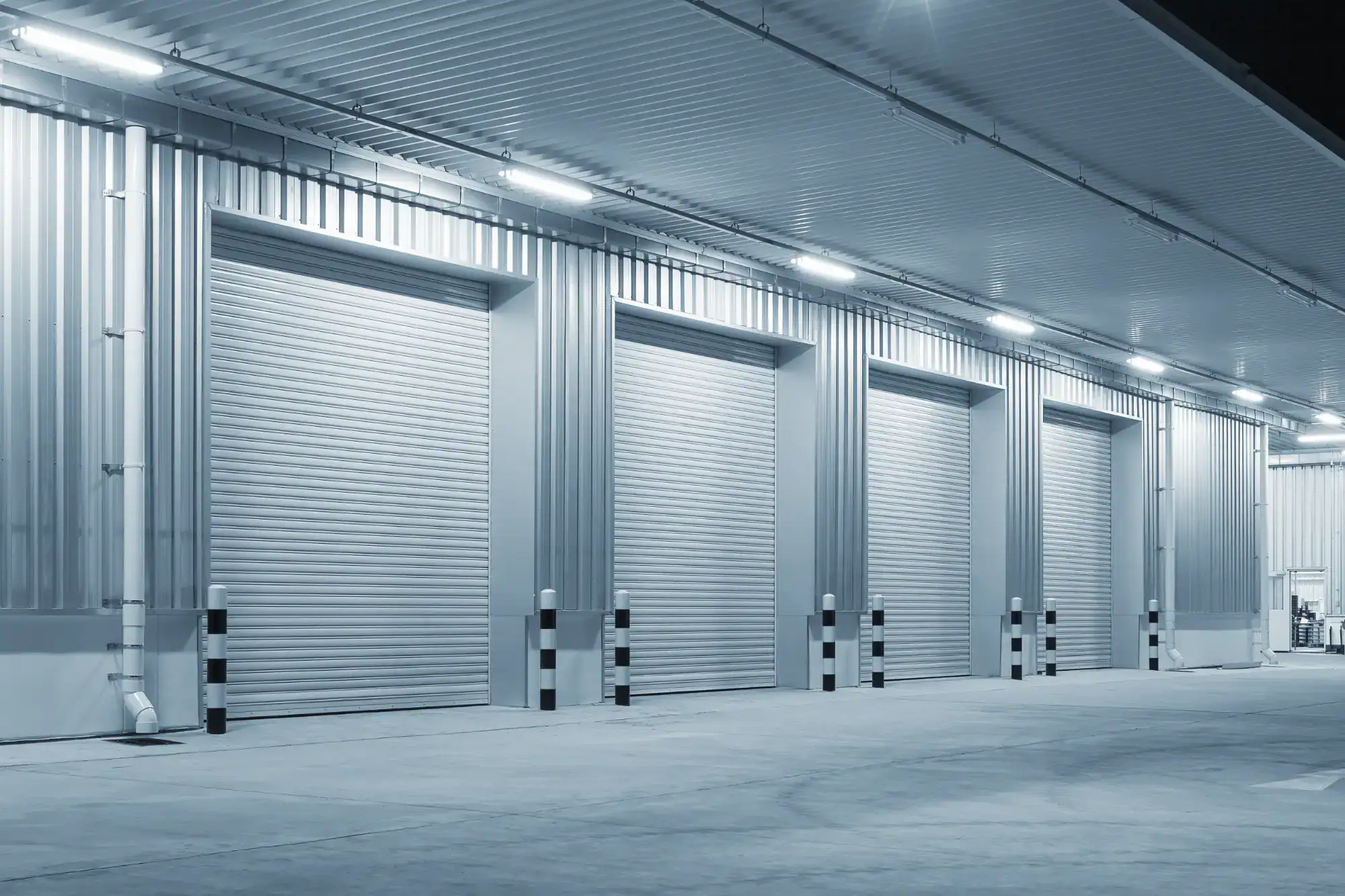 A row of large, closed metal garage doors on an industrial building at night, illuminated by overhead lights, with a concrete driveway in front.