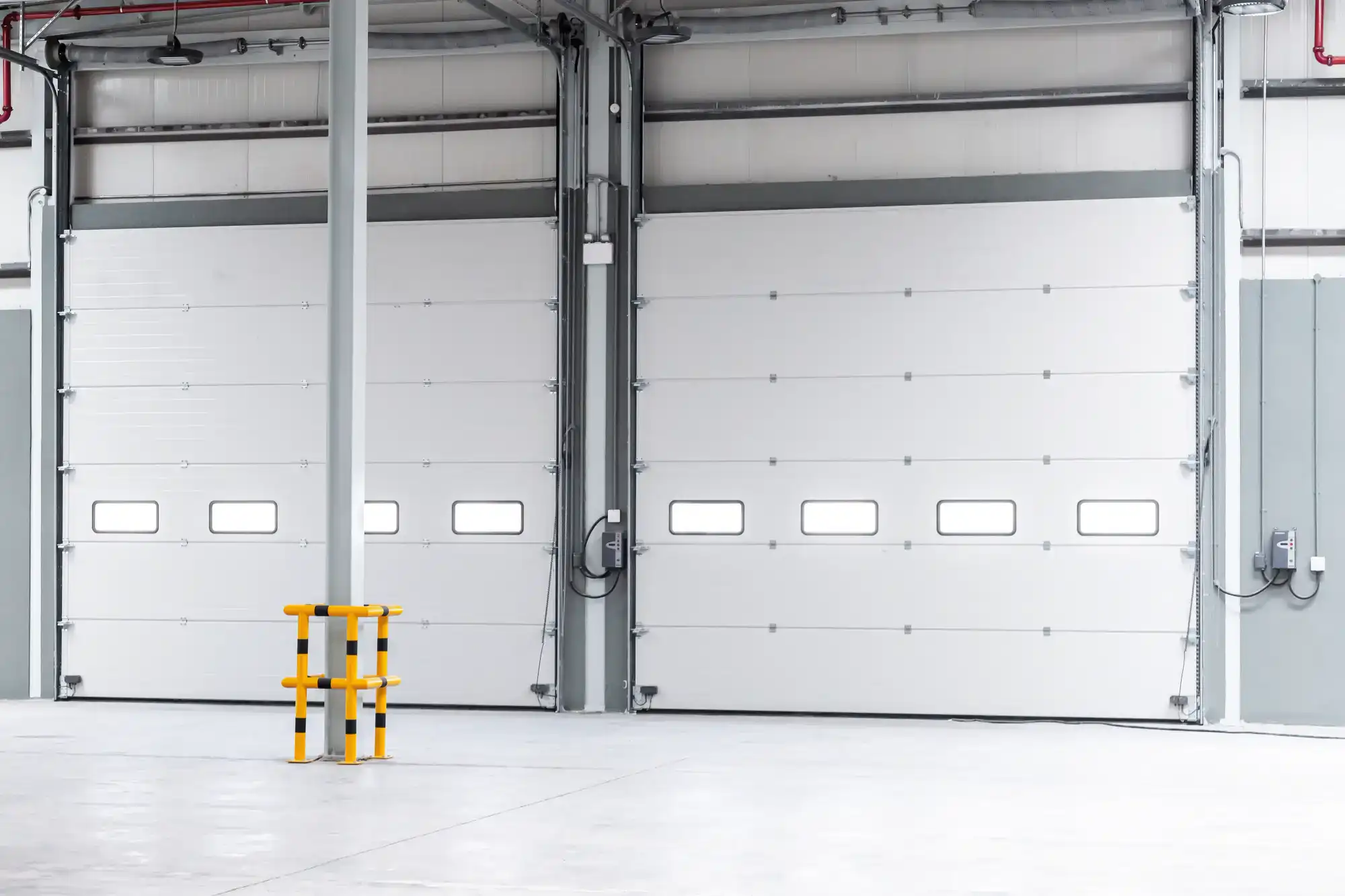 Two large white industrial garage doors with small rectangular windows near the bottom, located inside a clean, empty warehouse. A yellow safety barrier is positioned in front of a pole.