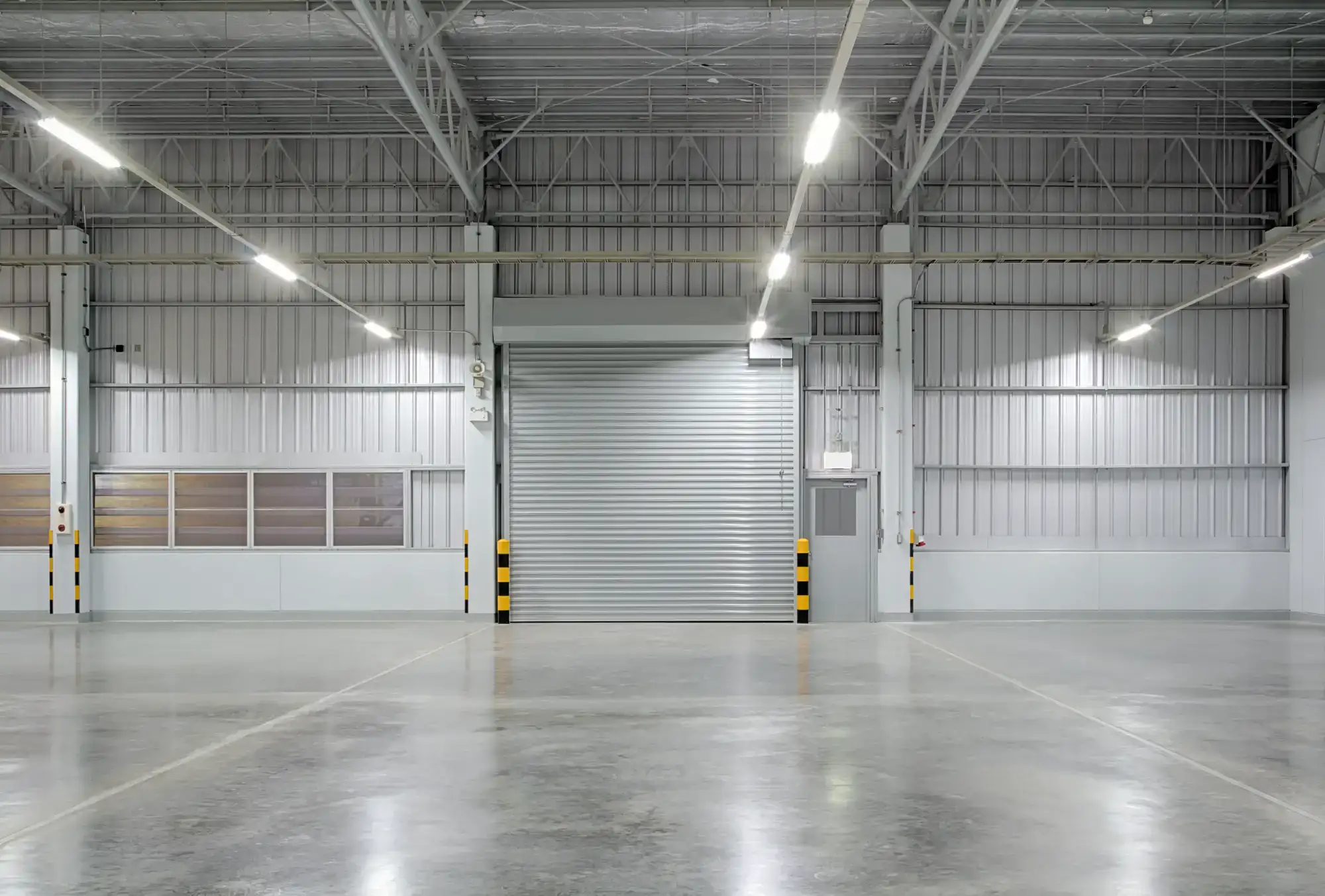 Empty, clean warehouse interior with polished concrete floors, high ceiling, fluorescent lighting, a large closed metal roll-up door, and white walls with exposed beams and pipes.