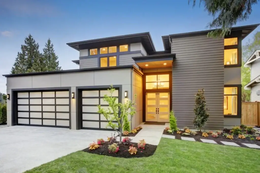 Modern two-story house with large windows, a three-car garage, and a well-maintained front yard featuring flowers and small trees, set against a clear sky with surrounding trees in Nassau County, NY.