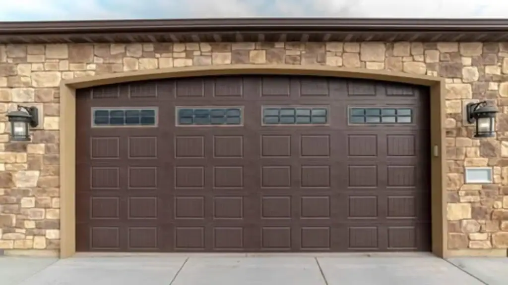 A closed brown garage door with rectangular patterns and small windows at the top, set in a wall made of tan and brown stone. Two black lantern-style lights are mounted on either side of the door in Nassau County, NY.