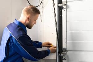 A man in a blue work uniform uses a screwdriver to adjust or repair the mechanism next to a white garage door, showcasing affordable garage door services inside a tidy garage.