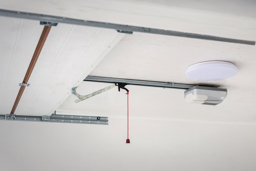 Close-up view of a garage door opener mechanism mounted on the ceiling, featuring a metal rail, a red emergency release cord, and a circular ceiling light.