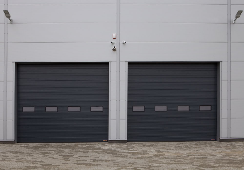 Two large, dark gray industrial garage doors with small rectangular windows, set in a light gray building wall above a paved surface. Security cameras and outdoor lights are mounted between and above the doors.
