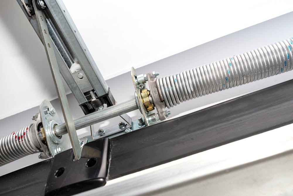 Close-up view of a garage door mechanism, showing metal torsion springs, a steel shaft, and mounting hardware attached to a horizontal black metal beam.