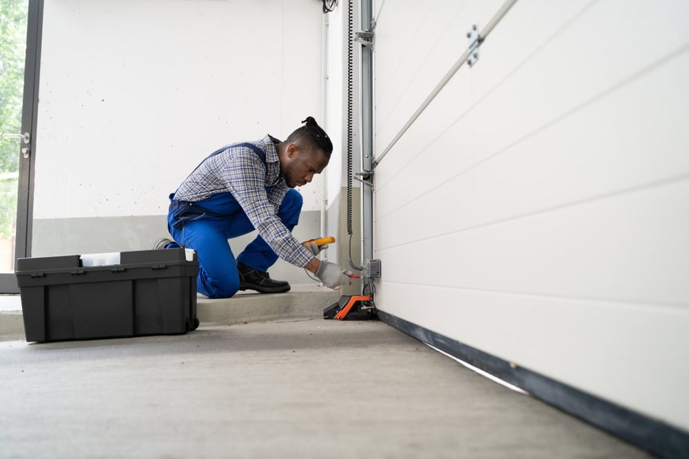 A man wearing blue overalls kneels on the ground, repairing a garage door with tools. A toolbox is open beside him as he works near the bottom mechanism, showcasing affordable garage door solutions in action.