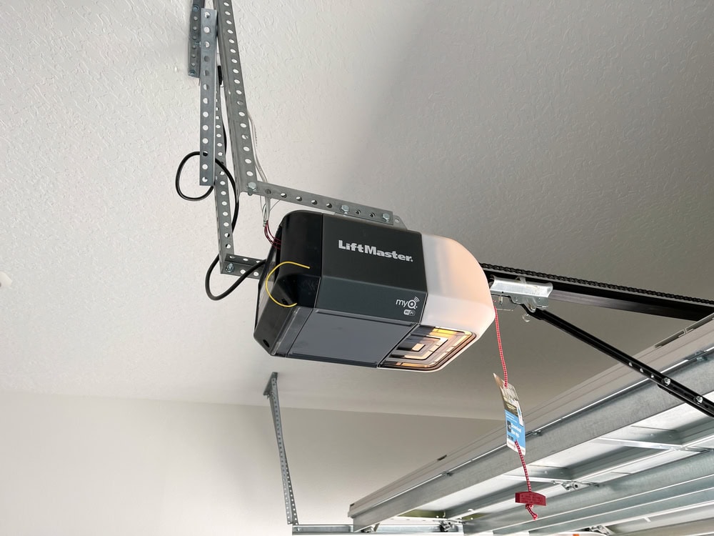 A LiftMaster garage door opener is mounted to the ceiling with metal brackets. The opener is attached to a partially open garage door, and a red emergency release cord hangs down.