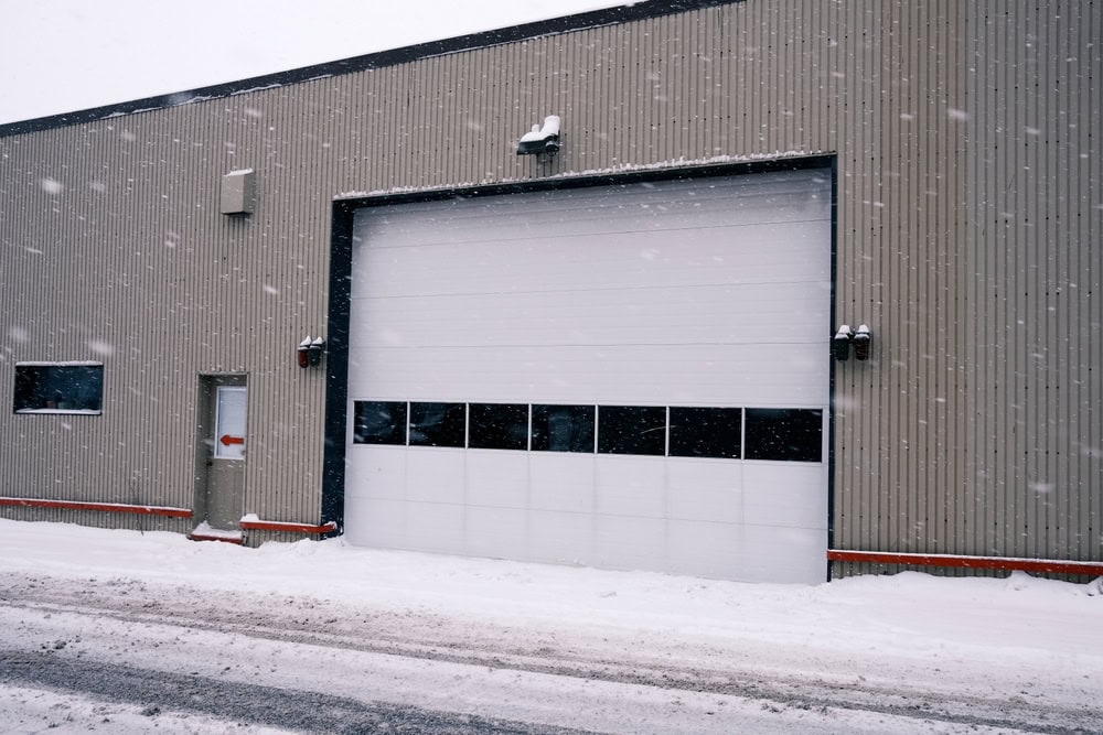 A large industrial garage door and a small entry door on the side of a beige building, with snow falling and accumulated on the ground and roof.