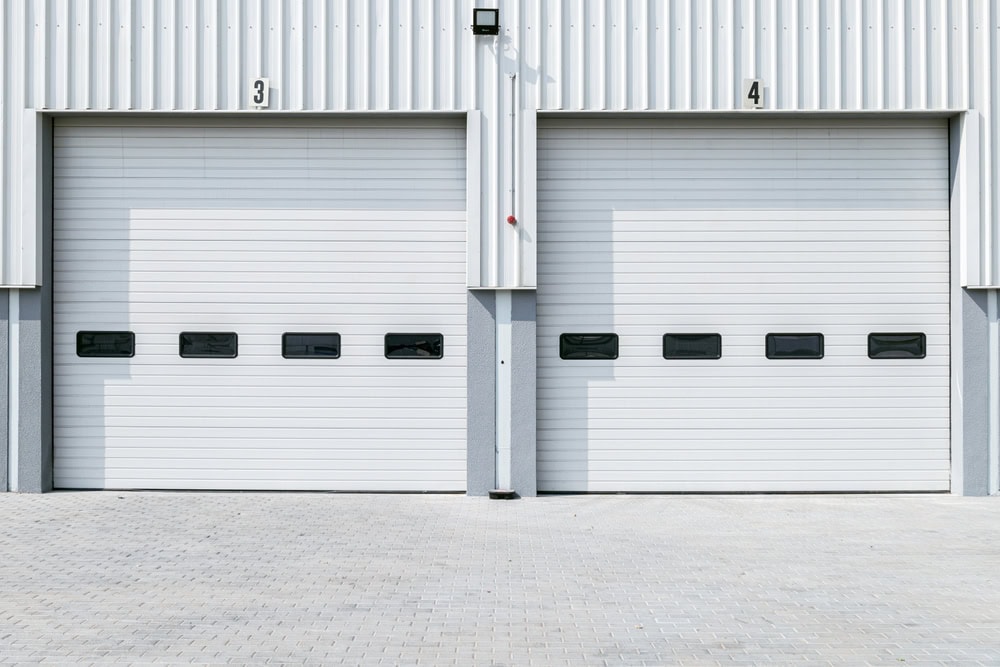 Two large white industrial garage doors, labeled 3 and 4, are closed. Each door has four rectangular windows in a horizontal row. The ground is paved with light gray bricks.