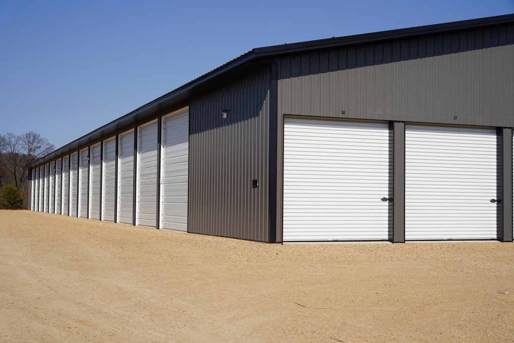 A row of metal storage units with white roll-up doors and a gravel driveway, photographed under a clear blue sky.