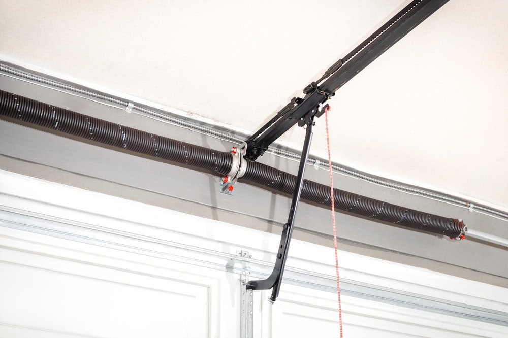 Close-up view of a garage door torsion spring system mounted above a white garage door, showing the metal shaft, spring, cables, and opener mechanism with a red emergency release cord.
