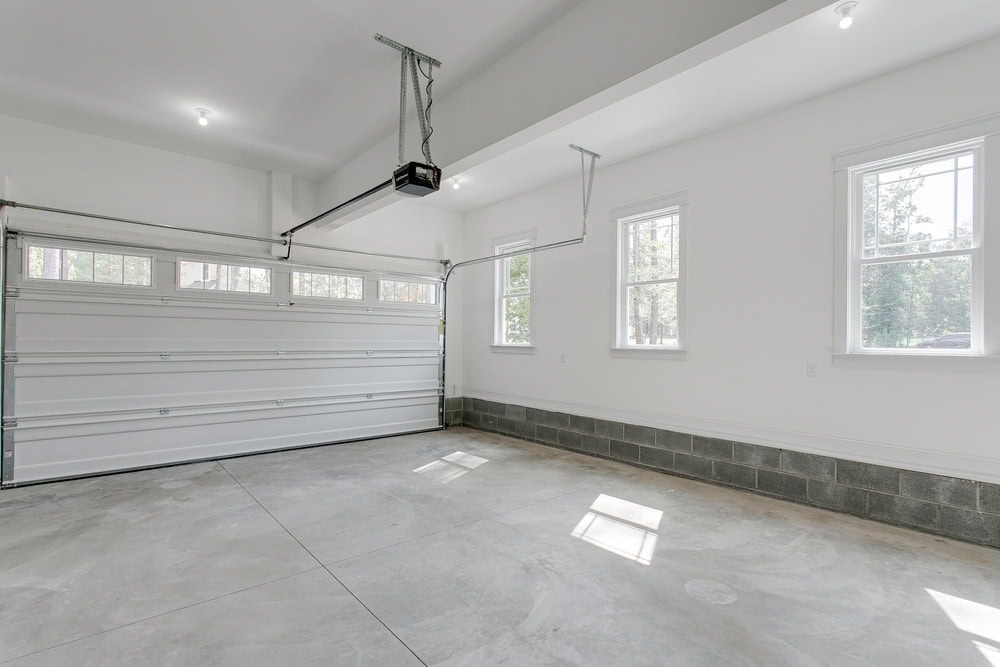 A clean, empty two-car garage with a closed white automatic garage door, white walls, large windows letting in natural light, and a smooth concrete floor.