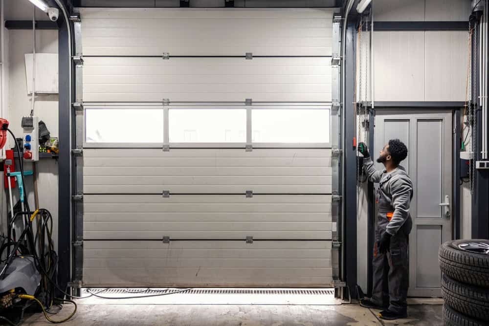 A person in work clothes stands near a wall, pressing a button to operate a large, closed garage door inside an industrial setting offering affordable garage door solutions. Equipment and tools are visible throughout the space.