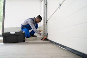 A man in blue overalls kneels on a garage floor, repairing a garage door with tools. A large toolbox is open next to him, showing affordable garage door services in action as the partially closed door is expertly fixed.