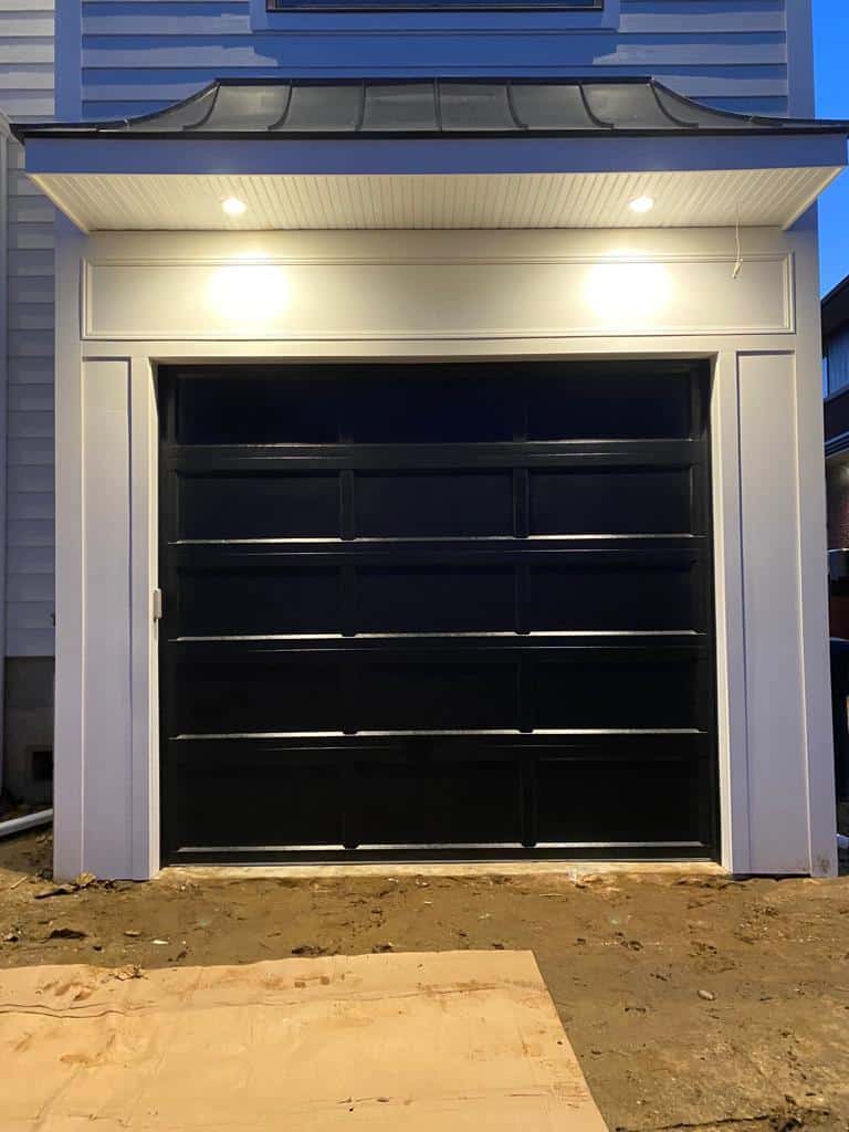 A black garage door with a modern design, illuminated by two overhead lights, set in a white frame on a house exterior. The ground in front appears unfinished, offering convenient garage access despite visible dirt and a plywood board.